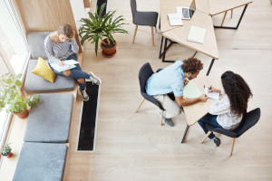 Two young startupers sitting at table at coworking space talking about team project, looking through information. Girl sitting on window sill, getting ready for exams